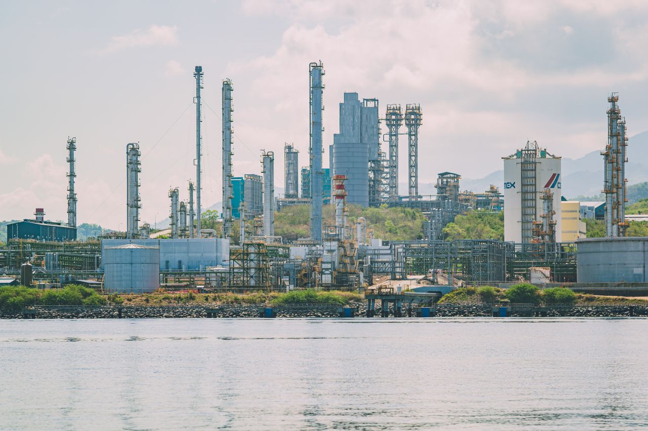 A large industrial refinery complex with tall chimneys by the waterfront under a clear sky.